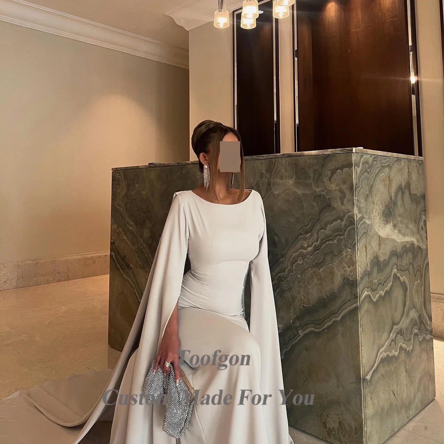 Woman in an elegant white gown with long sleeves, holding a silver clutch. She stands beside a marble counter under modern pendant lights in a neutral-toned room.