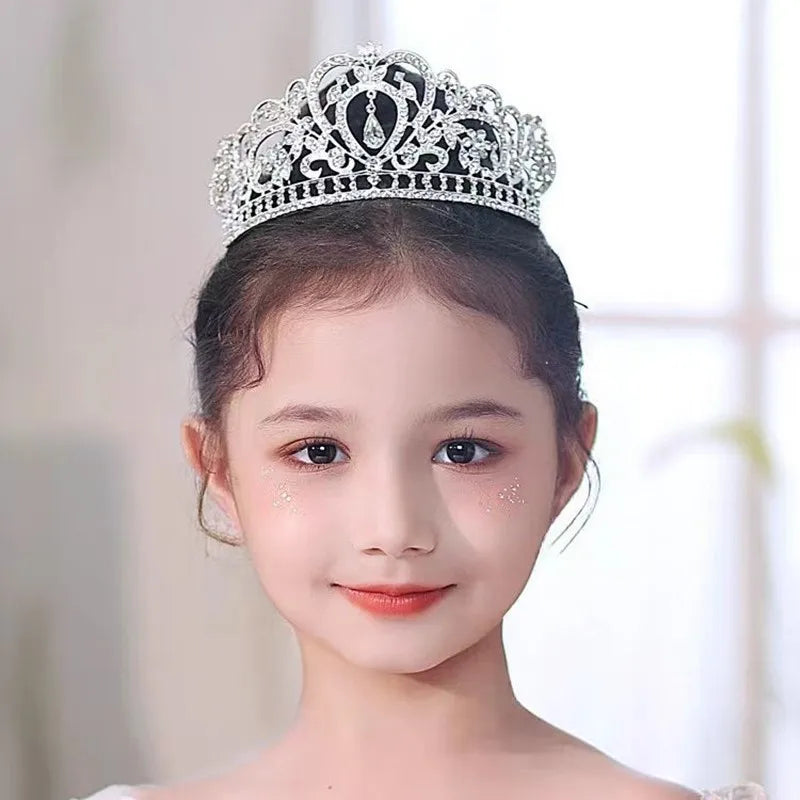 Young girl with a jeweled tiara and sparkly makeup smiles softly against a blurred light background, highlighting her delicate features and elegant attire.