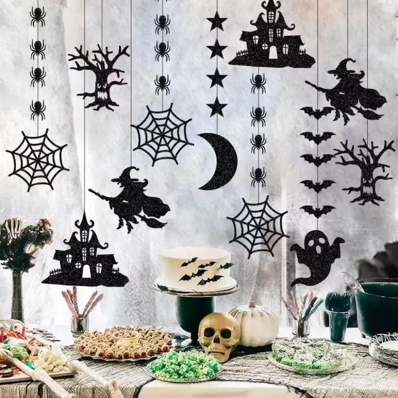 Halloween-themed table setting with black hanging decorations featuring witches, haunted houses, spiders, cobwebs, bats, and ghosts. The table is adorned with skulls, festive treats, a cake with bat designs, candy bowls, and white and green pumpkins, creating a spooky atmosphere.