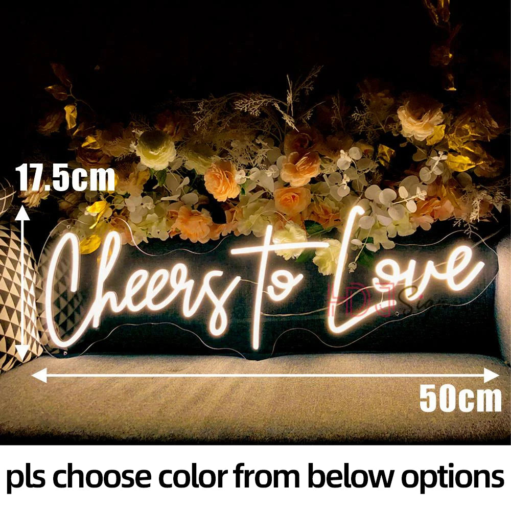 Neon sign reading "Cheers to Love" in cursive, measuring 17.5cm by 50cm, set against a backdrop of flowers and a patterned pillow, with a color selection note below.
