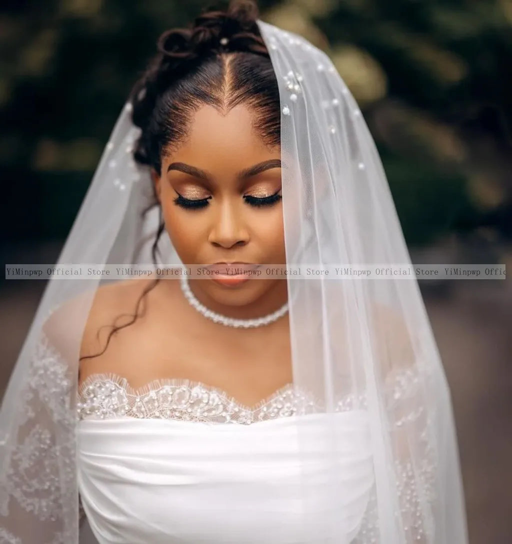 A bride in a white lace gown gently smiles, her eyes closed under a pearl-embellished veil. Her hair is elegantly styled, set against a blurred outdoor background.