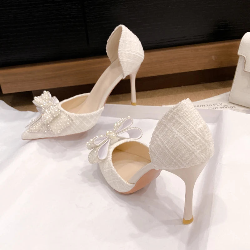 Elegant white high heels with textured fabric, adorned with pearl-embellished bows on the toes. The shoes rest on a white surface against a wooden background.