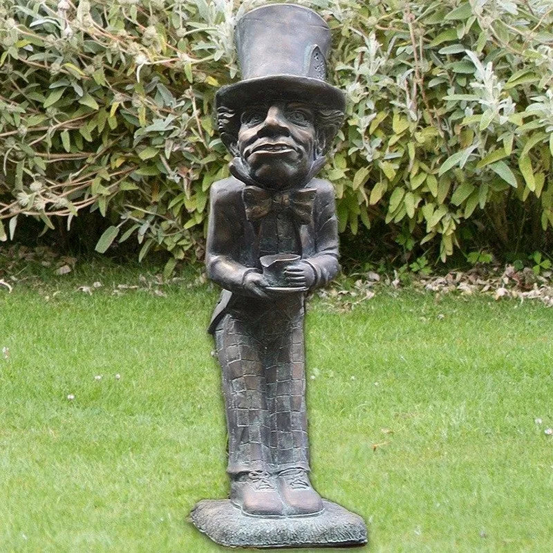 A whimsical bronze statue of a tall, thin man in a top hat and bowtie, standing in a garden with lush green grass and leafy bushes in the background.