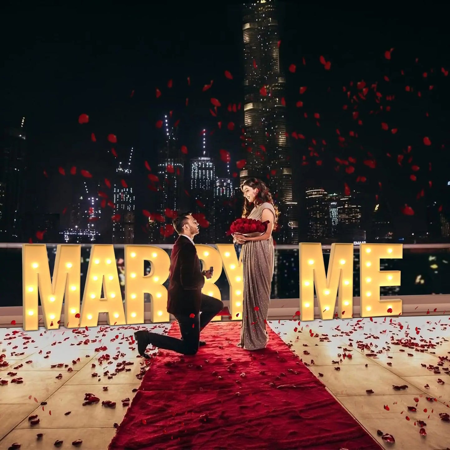 Man kneels, proposing with roses to a woman on a rooftop adorned with a red carpet and rose petals. Illuminated "Marry Me" sign and city skyline backdrop complete the scene.