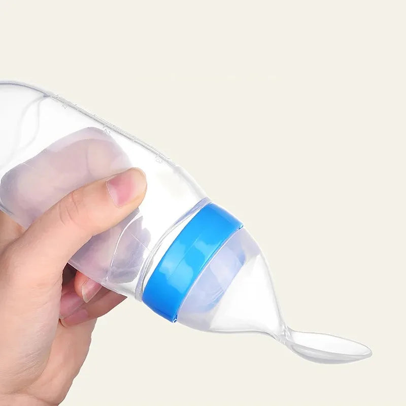 A hand holding a clear plastic baby bottle with a blue cap, inverted, showing a built-in spoon at the tip against a neutral background.