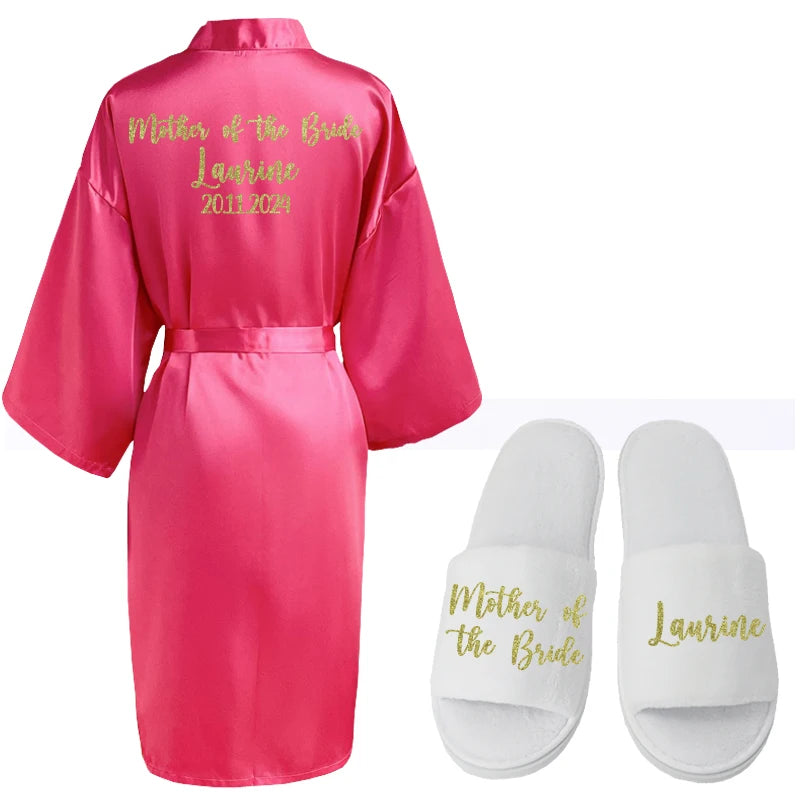 A pink satin robe with "Mother of the Bride," a name, and years embroidered in gold, paired with white slippers bearing matching text.