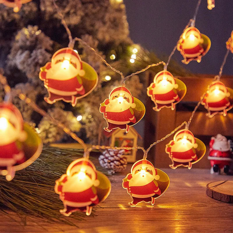 Festive string lights featuring tiny Santa Claus figures illuminate a cozy Christmas scene with a snowy tree and pinecones in the background, creating a warm holiday ambiance.