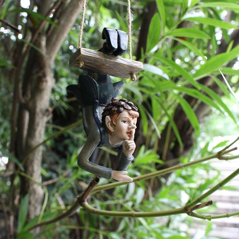 Hanging garden ornament of a fairy with pointed ears, resting on a wooden swing amid green foliage. The fairy is posed with a finger to its lips, signaling silence.