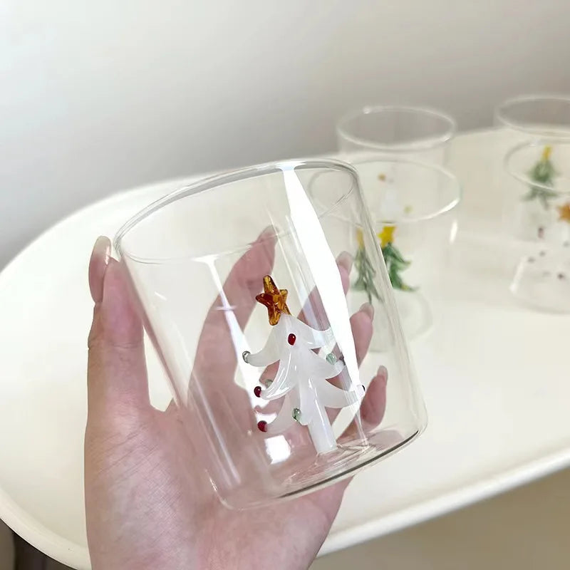 Hand holding a clear glass with a white Christmas tree design, topped by a golden star, against a blurred background of additional similar glasses on a white surface.