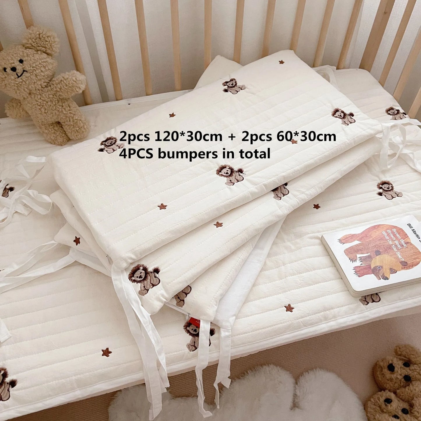 Pile of white crib bumpers with bear and star patterns, totaling four pieces. Displayed on a crib with a teddy bear and a children's book nearby.