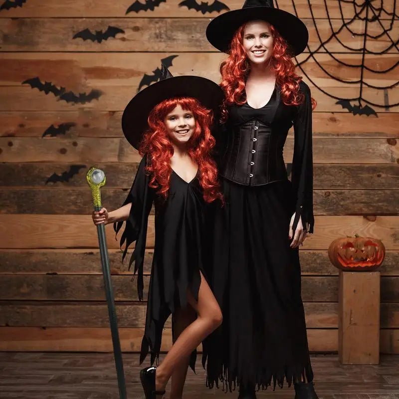 Two people in matching witch costumes stand indoors. They wear black dresses, red wigs, and hats. The backdrop features bats and a carved pumpkin on a wooden shelf.
