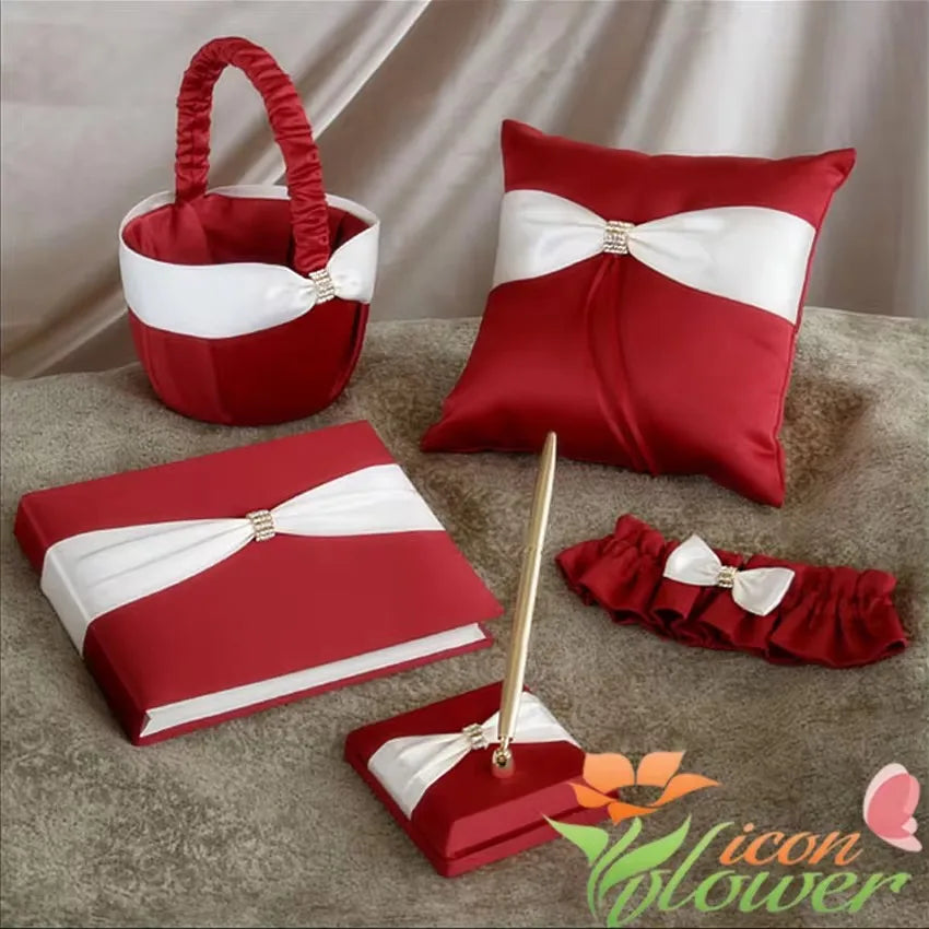 A red and white wedding set includes a pillow, basket, guest book, pen holder, and garter, all adorned with white ribbons and rhinestone accents on a soft surface.