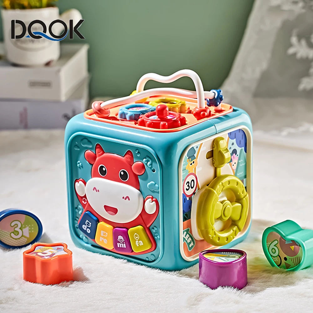 Colorful educational toy cube with animal illustrations, interactive buttons, and musical symbols. Placed on a soft surface with small shape pieces scattered around.