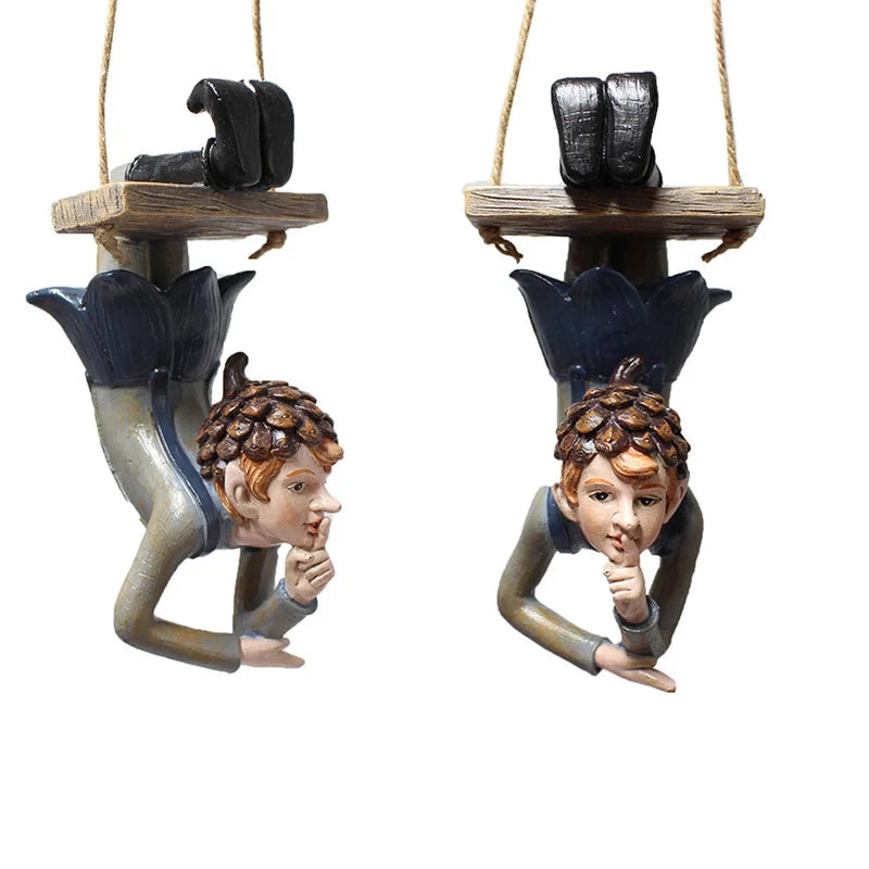Whimsical figurines of mischievous elves hanging upside down from wooden swings, with finger pressed to lips, brown acorn caps, and blue flower bud bodies.