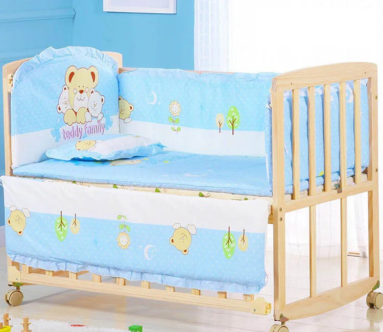 Baby crib with light wood frame, blue bedding featuring cute bear and tree designs. Side guard has cartoon image with "Teddy family." Crib is on wheels, set in a bright room.