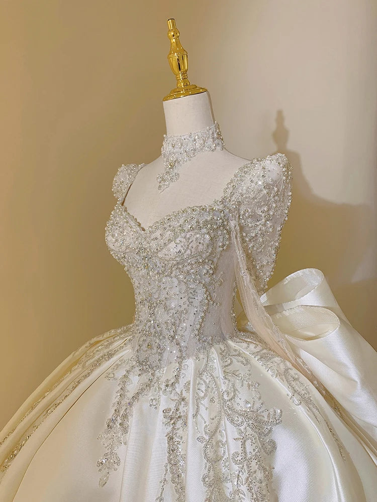 Elegant white gown on a mannequin, featuring intricate silver embroidery, beading on sheer long sleeves, a high neckline, and a voluminous skirt with floral patterns.