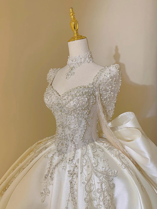 Elegant white gown on a mannequin, featuring intricate silver embroidery, beading on sheer long sleeves, a high neckline, and a voluminous skirt with floral patterns.