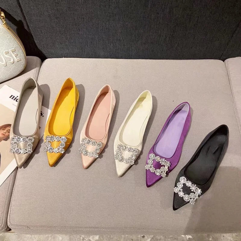 Six pairs of jeweled, pointed-toe flats in various colors—beige, yellow, pink, white, purple, and black—are arranged on a light gray sofa.