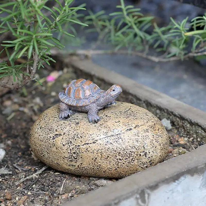 A small tortoise figurine with an orange and gray shell rests on a tan speckled stone amidst soil and green plants in a garden setting.