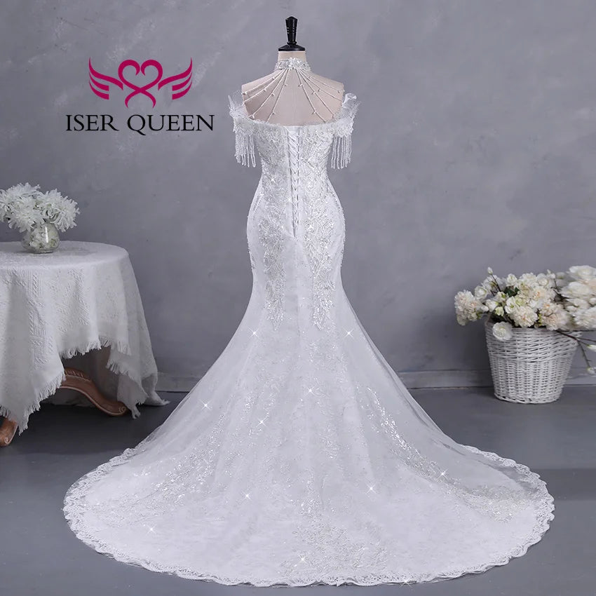 Elegant white bridal gown with off-shoulder beaded sleeves, displayed on a mannequin. Features a long lace train. Flowers in vase and basket in the background.