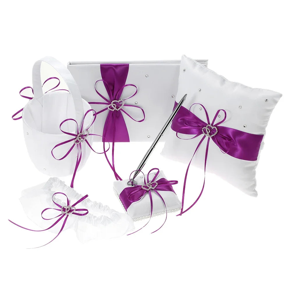 White wedding accessory set with purple satin ribbons and rhinestone hearts. Includes a pillow, basket, garter, guest book, and pen, all adorned with decorative bows.