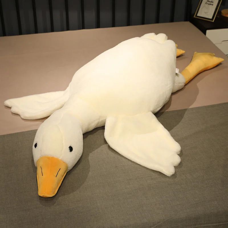 A large, soft white goose plush toy with an orange beak and feet lies sprawled on a bed, creating a cozy and inviting atmosphere in the room.
