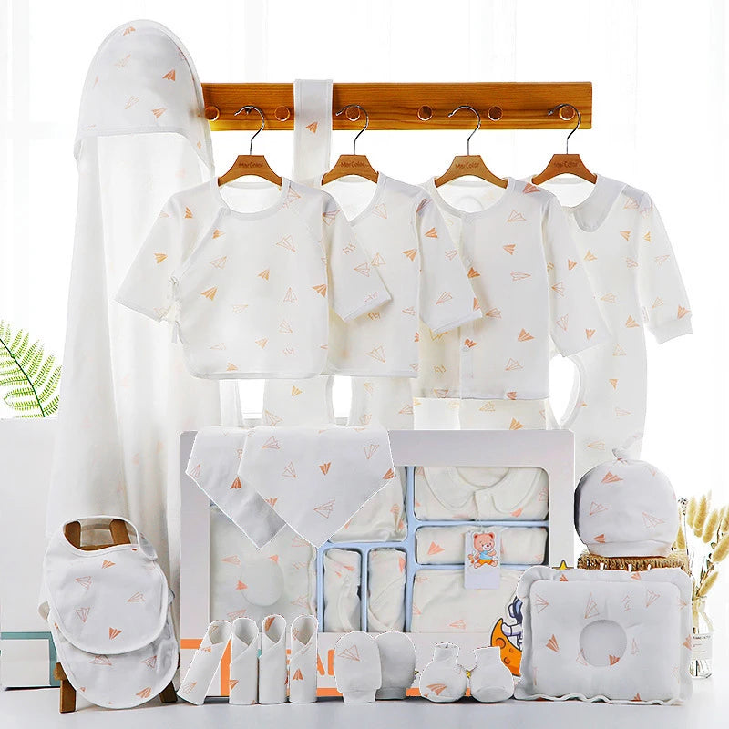 White baby clothing set with paper airplane prints. Includes shirts, bibs, hats, mittens, socks, and a pillow, all displayed on a wooden rack with soft lighting.
