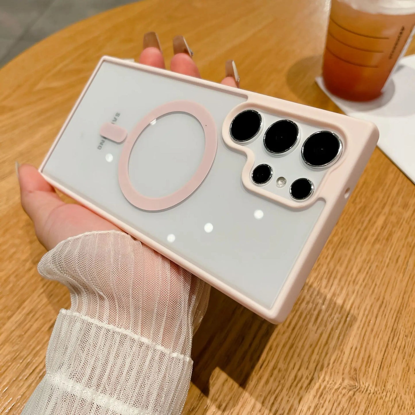 A hand with long nails holds a smartphone with a pink and white case featuring multiple camera lenses on the back. The phone is held over a wooden table with a drink in the background.