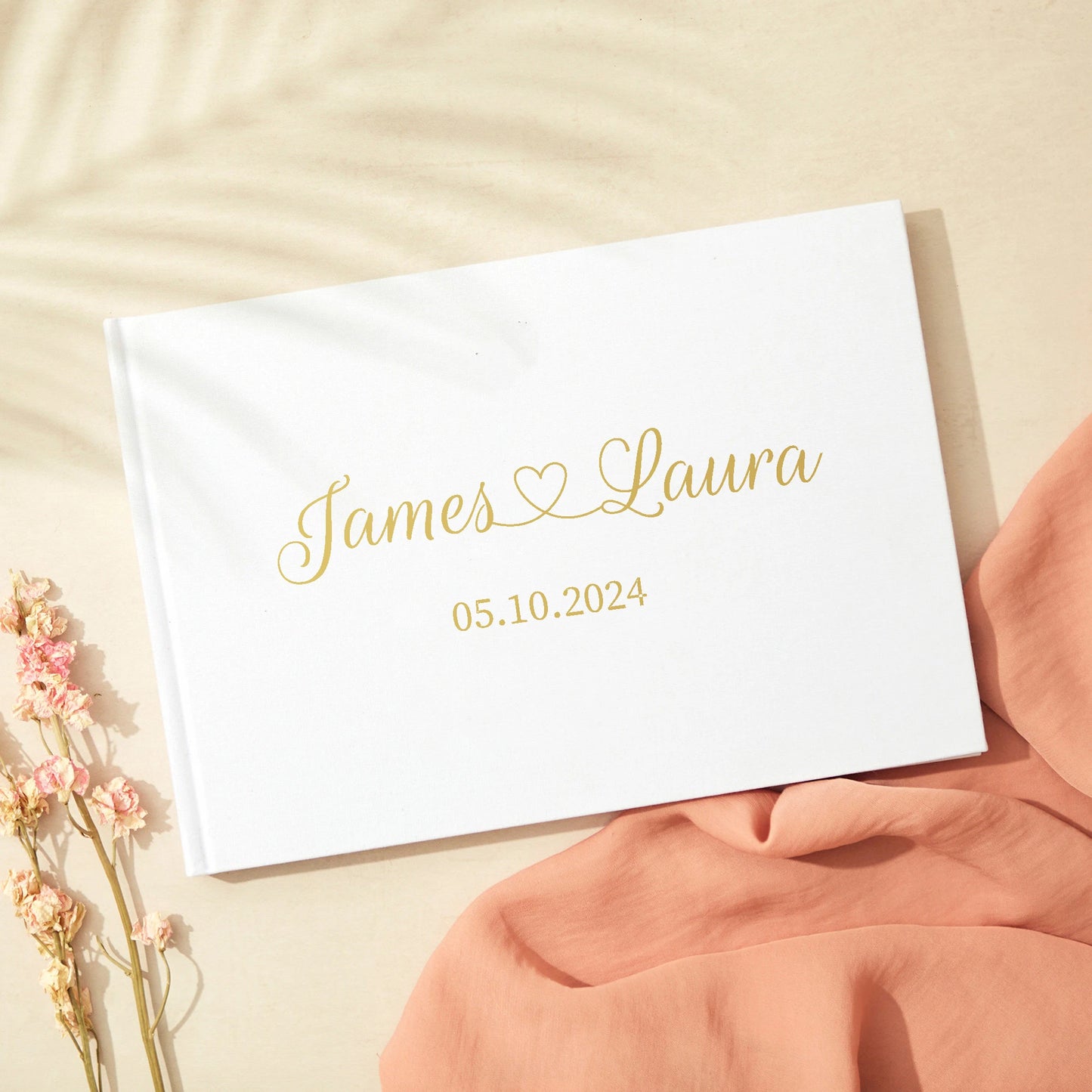 Wedding guest book with "James ♥ Laura" and date "05.10.2024" in gold script on a white cover, next to pink flowers and fabric on a light surface.