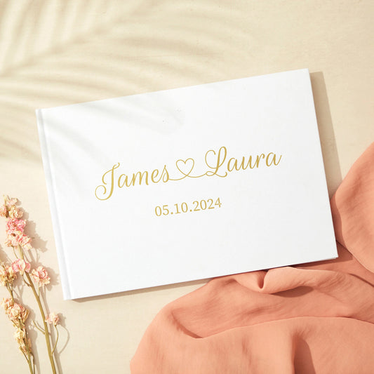 Wedding guest book with "James ♥ Laura" and date "05.10.2024" in gold script on a white cover, next to pink flowers and fabric on a light surface.
