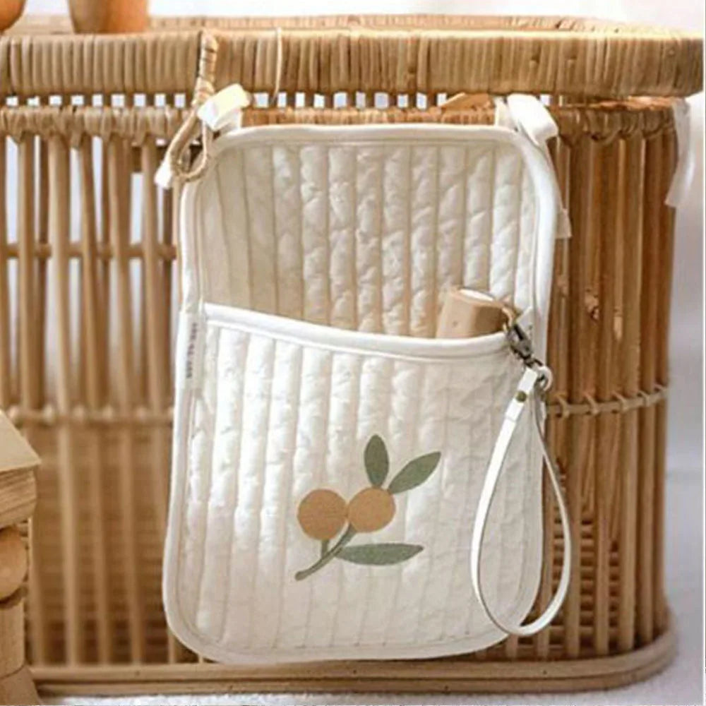 Quilted white bag with an orange fruit and leaf design, hanging on a wicker basket. A wrist strap is attached, and a wooden item is partially visible inside the pocket.