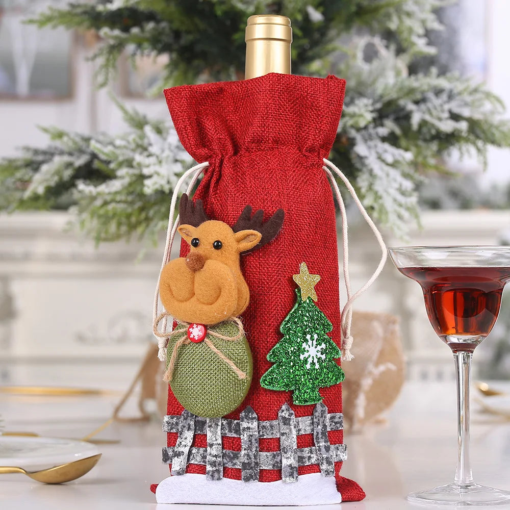 A festive wine bottle wrapped in a red bag with a reindeer and Christmas tree design, next to a filled wine glass, set against a snowy decorated tree.