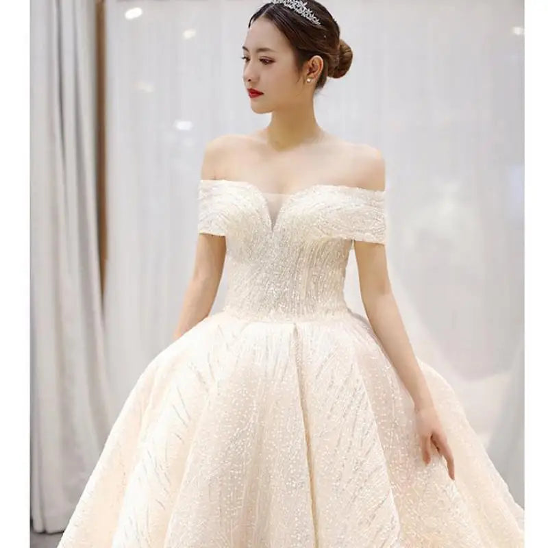 Woman in an off-shoulder, elegant white gown with a full skirt and textured fabric, wearing a tiara, stands against a soft, blurred background, looking to the side.