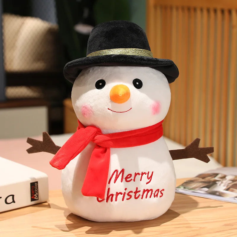 Plush snowman with a black hat, orange nose, and red scarf reading "Merry Christmas." It has stick-like arms and is sitting on a wooden surface beside a book.