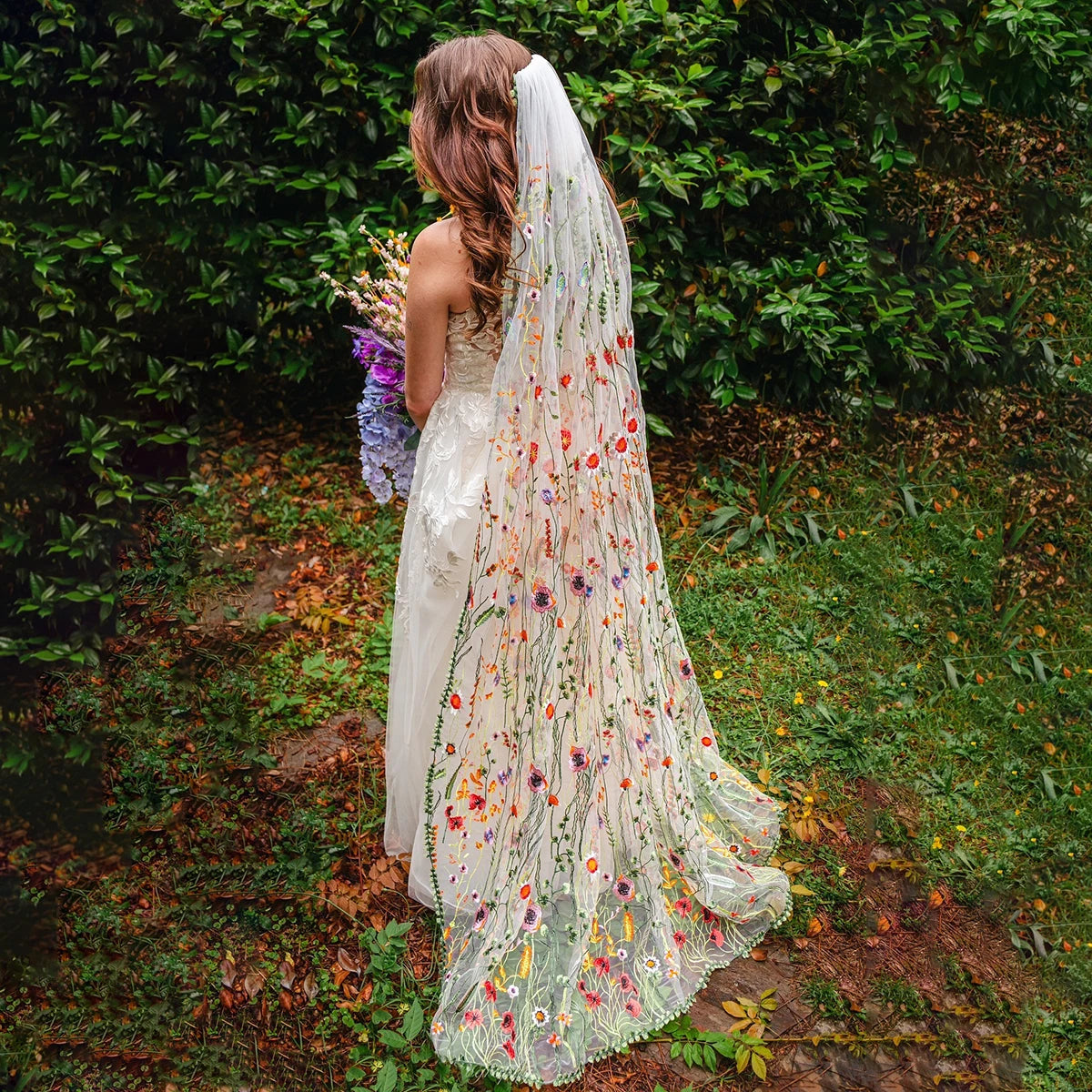 A bride stands in a garden, wearing a gown with an embroidered floral veil and holding a bouquet of purple and white flowers. Lush greenery surrounds her.