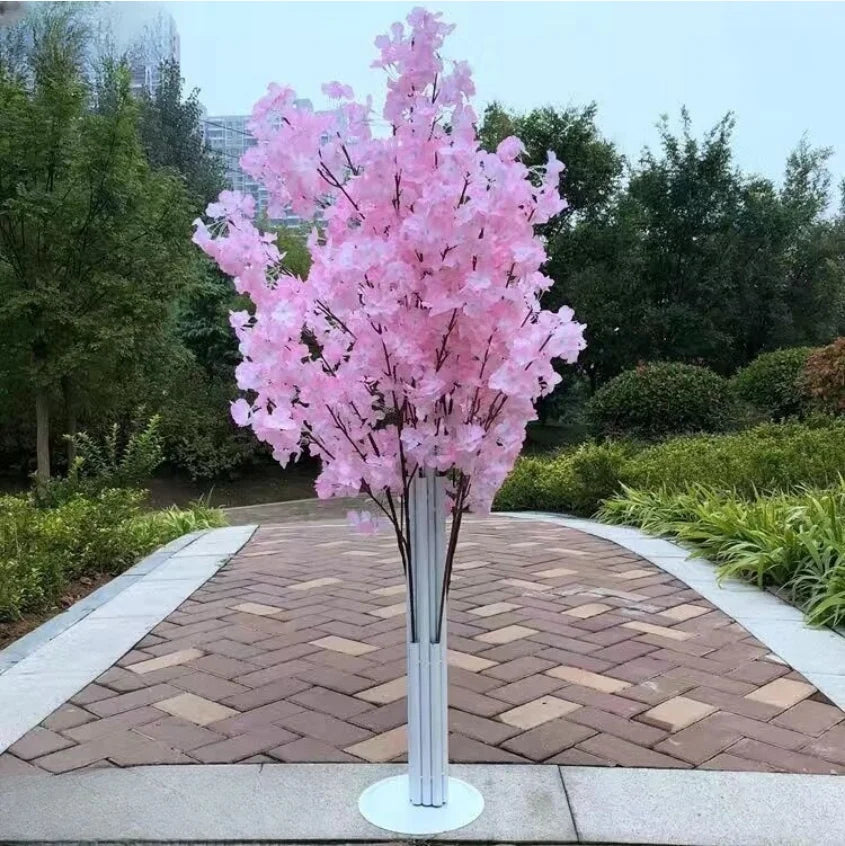 Artificial cherry blossom tree with pink flowers on a white stand, placed on a paved path surrounded by greenery and shrubs in an outdoor garden setting.