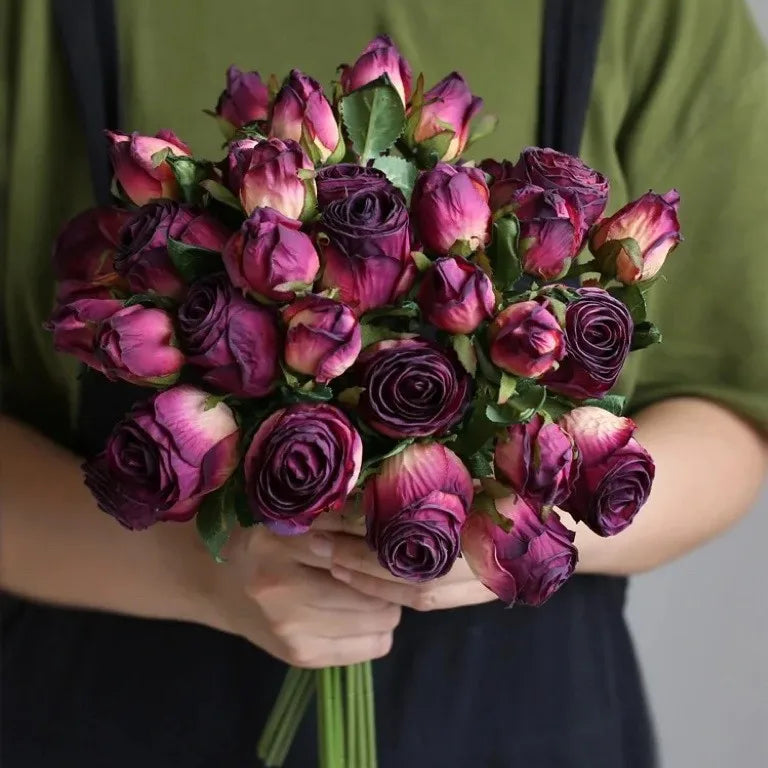 A person in a green and black outfit holding a bouquet of tightly clustered purple roses with hints of pink and green leaves, against a neutral background.