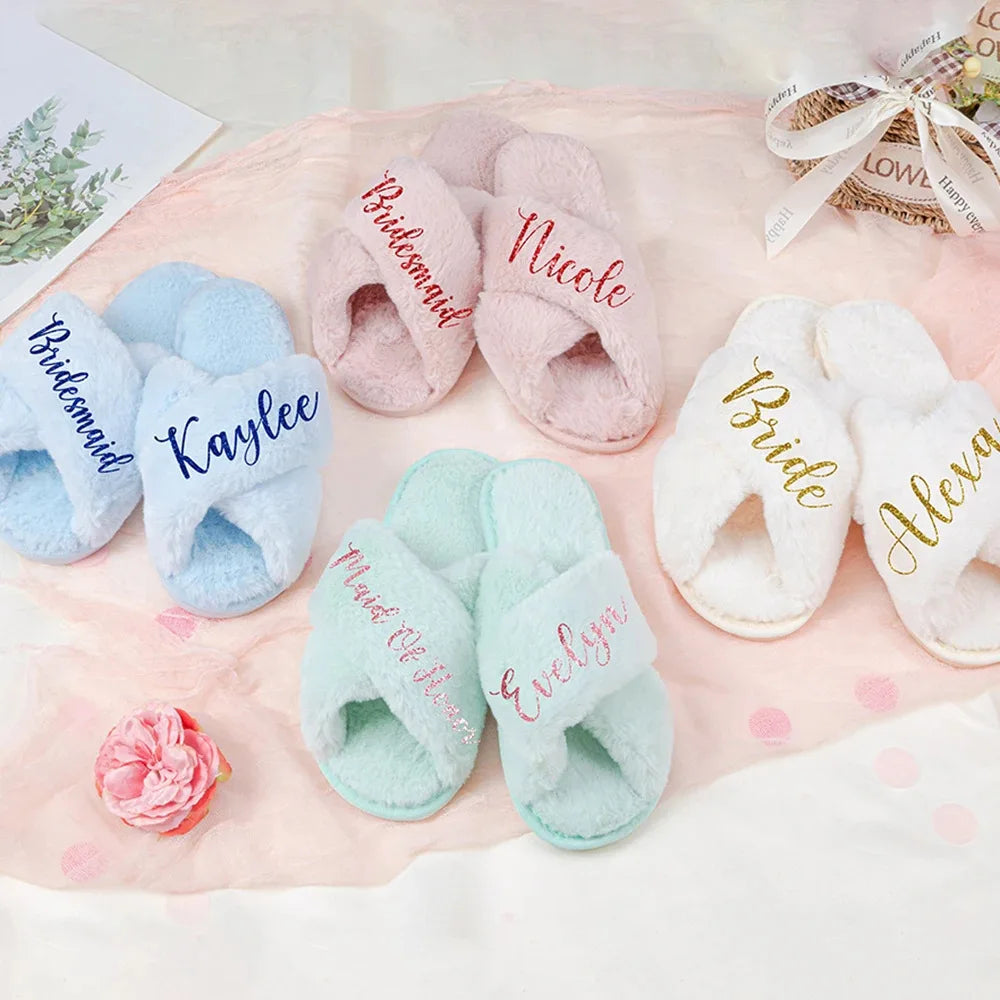 Cozy slippers in pastel pink, blue, mint, and white with cursive names like “Bride,” “Bridesmaid,” and “Kaylee.” Arranged on a soft blush fabric with a pink flower nearby.