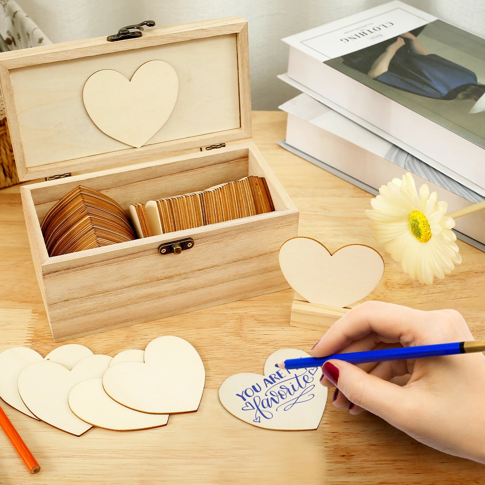 Wooden box with heart-shaped pieces, one being written on with "You are a favorite" using a blue pen. Books and a daisy are in the background on a wooden table.