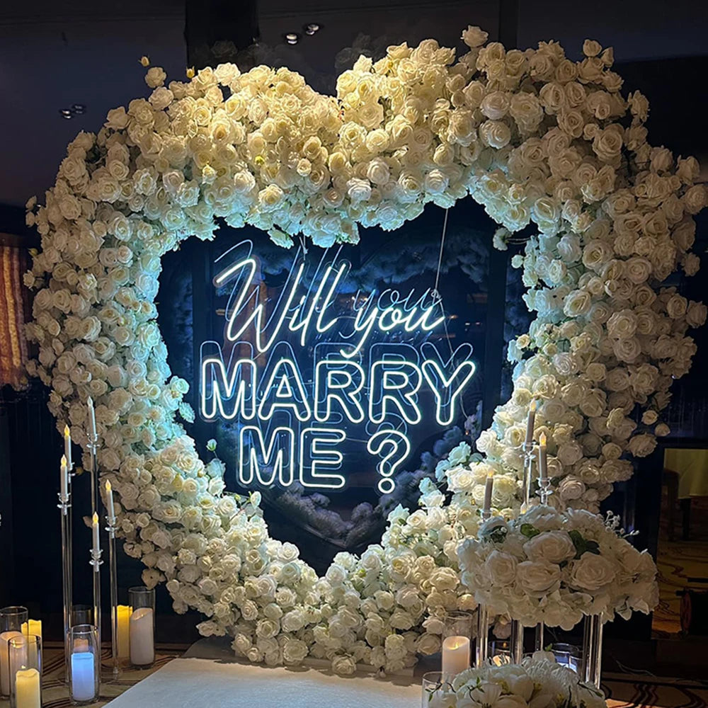 Heart-shaped floral arch made of white roses with a neon sign in the center reading "Will you marry me?" Surrounded by candles and elegant decor.