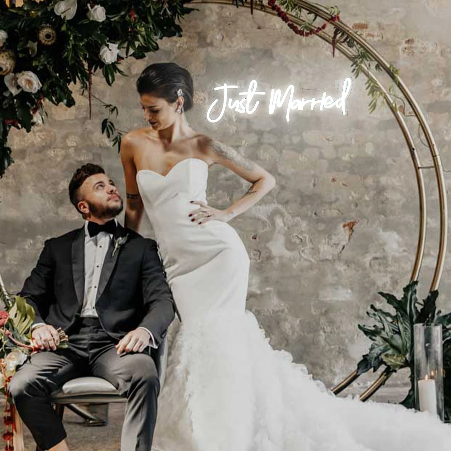 Bride in a strapless gown stands next to the seated groom in a tuxedo, under an ornate floral arch. "Just Married" glows on the wall, creating a romantic scene.