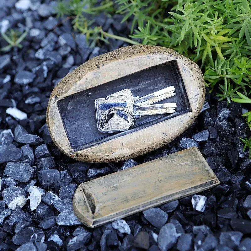 Hidden key rock with a removable lid on black gravel, surrounded by green plants. The key compartment is open, revealing silver keys inside.