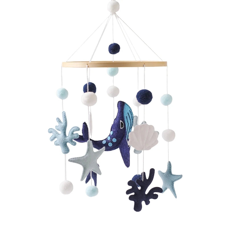 A felt baby mobile with a marine theme features a blue whale, starfish, seashell, and coral shapes, suspended with white and blue pom-poms from a circular wooden frame.