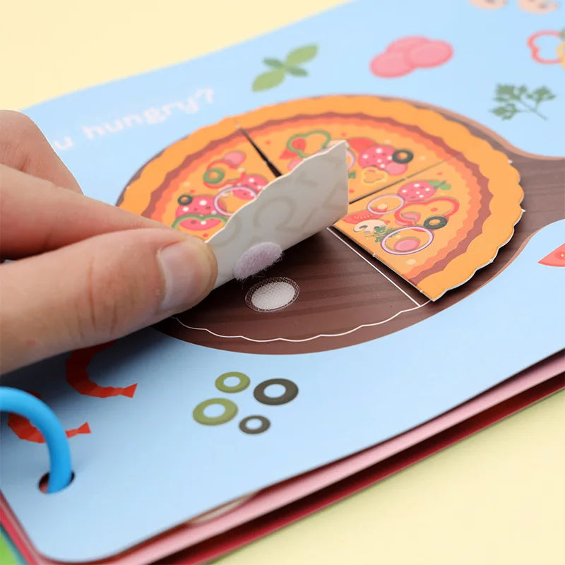 A hand lifts a slice of illustrated pizza from a children's book page, revealing a Velcro attachment. The page features colorful graphics and text reading "Are you hungry?"