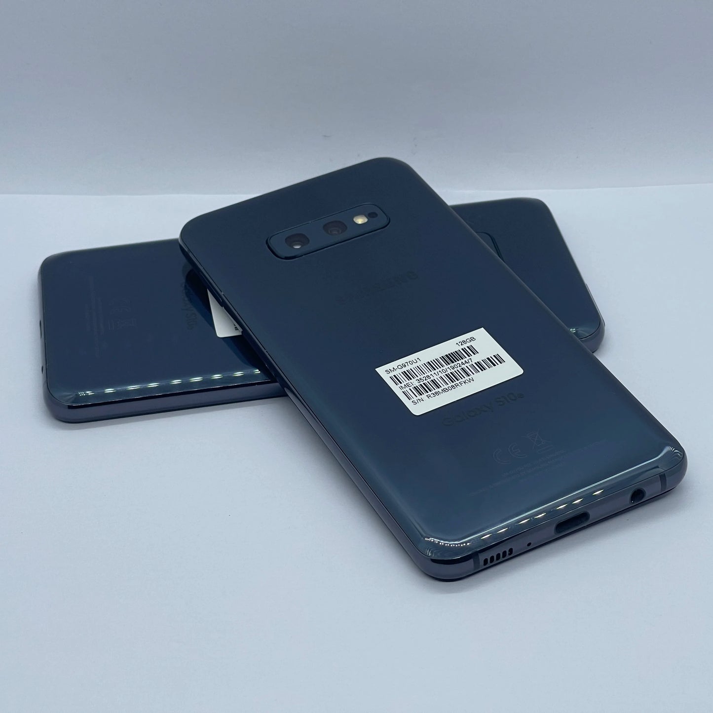 Three dark-colored smartphones stacked on a white surface, with visible dual rear cameras and a barcode sticker on the top phone.