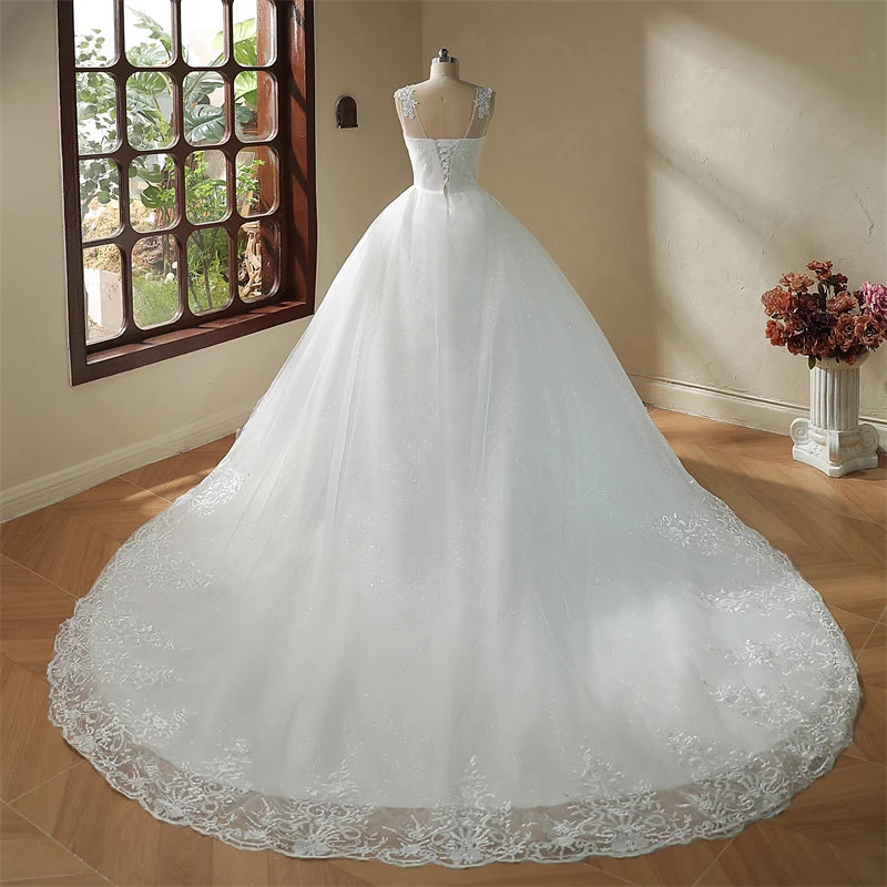 Elegant white wedding dress on mannequin by a window, featuring a fitted bodice, lace details, and a voluminous train with floral patterns. A vase of dried flowers is nearby.
