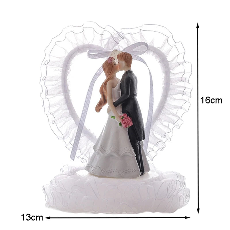 Bride and groom cake topper with a lace heart backdrop, measuring 16cm tall and 13cm wide. The bride holds a pink bouquet as the couple embraces on a ruffled base.