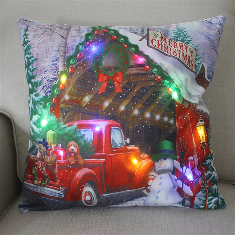 Festive pillow with a winter scene featuring a red truck carrying gifts and a dog, adorned with a wreath, snowman, and a "Merry Christmas" sign, illuminated by colorful lights.