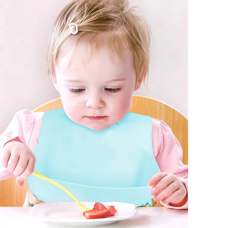 A baby wearing a blue bib sits in a high chair, focused on scooping sliced strawberries from a plate with a yellow spoon. The baby has light hair with a small clip.