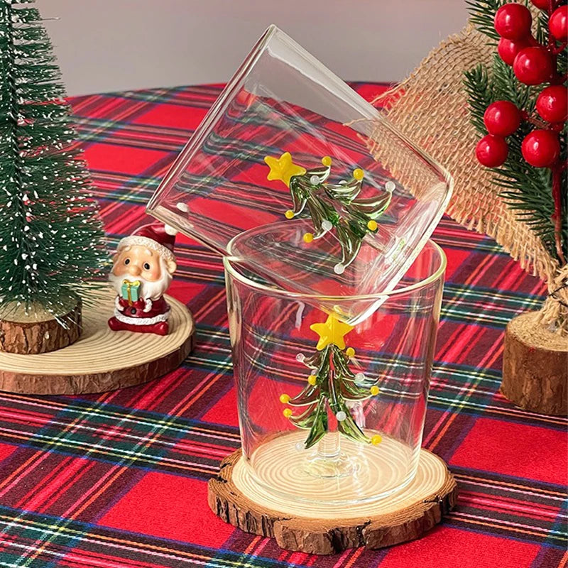 Two glass cups with colorful glass Christmas tree inserts on a plaid tablecloth, surrounded by a small decorative tree, Santa figurine, and red berry arrangement.